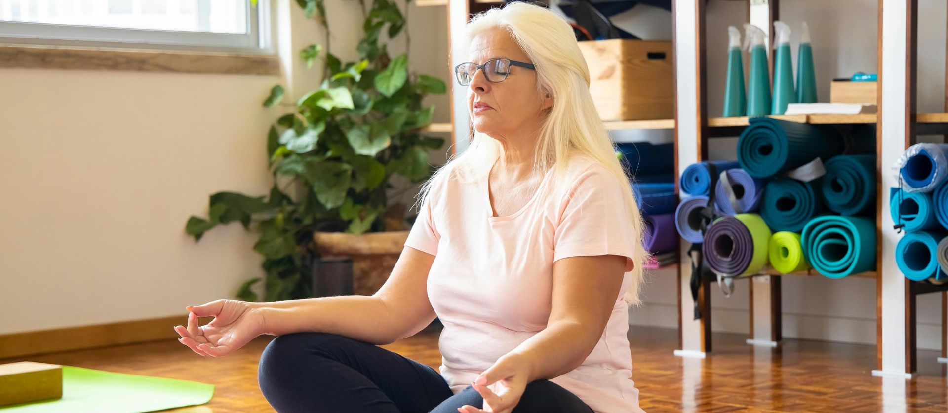 Adult woman sits cross-legged on a mat meditating in a serene yoga studio environment.