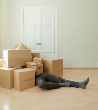 A person lying under a pile of moving boxes in a minimalistic room conveys tiredness from packing.