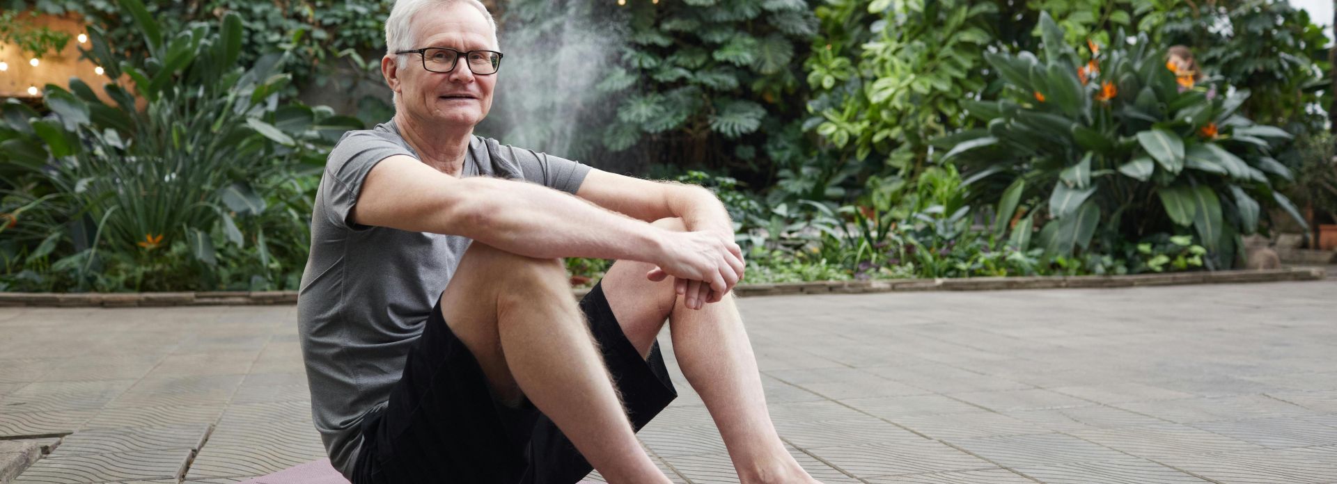 Older man enjoying yoga session on a mat in a lush botanical garden.
